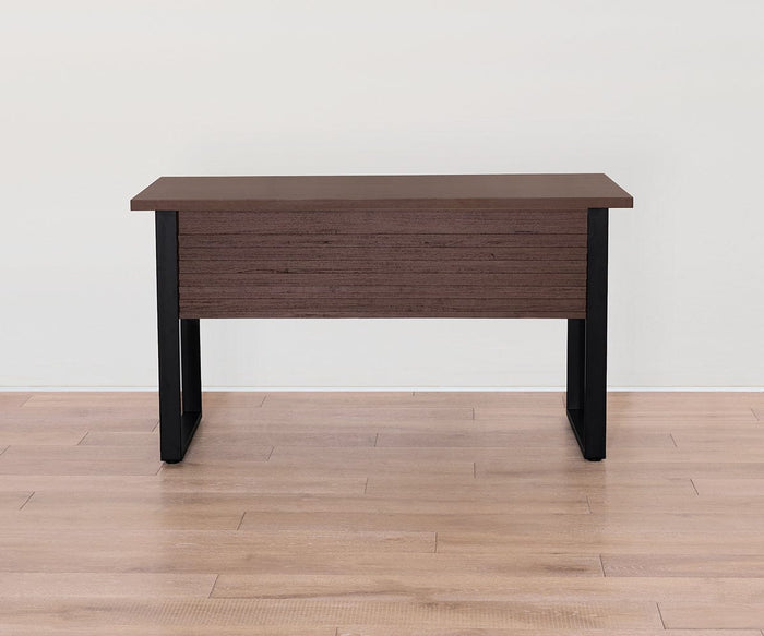 A sleek rectangular Cary Desk with a brown melamine top and black legs stands on a light wood floor against a plain white wall.