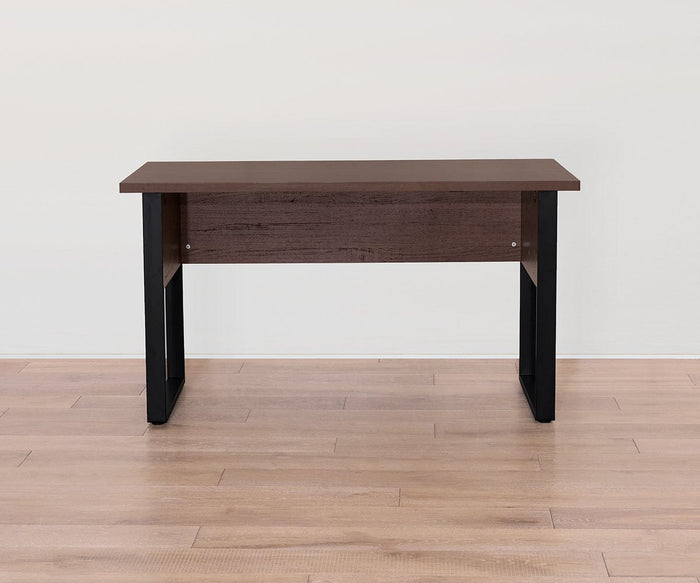 A minimalist Cary Desk with a 55″ dark brown walnut-finished top and black rectangular legs, set on a light wooden floor against a plain white wall.