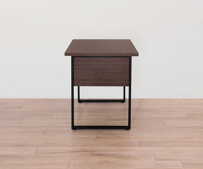 A Cary Desk with a 55″ melamine top in rich walnut finish and black metal legs is positioned on a light brown wooden floor against a plain white wall, viewed from the side.