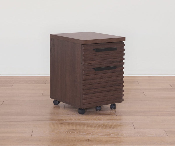 The Cary Mobile Pedestal, featuring a small profile with a rich walnut finish and two sleek black handles, rests on four caster wheels atop a light wooden floor against a plain white wall, providing practical office storage.