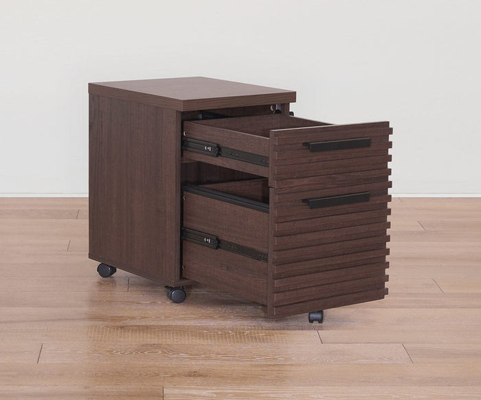 The Cary Mobile Pedestal, finished in rich walnut, features two drawers—including one open to showcase ample office storage space. This dark wood file cabinet is set on wheels and positioned on a light wooden floor against a plain, light-colored wall.