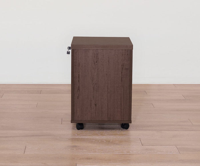 The Cary Mobile Pedestal, featuring a rich walnut finish and brown wooden construction, sits on wheels atop a light wood floor against a plain white wall. The side view displays a lock on the left, making this mobile pedestal an excellent choice for secure office storage.