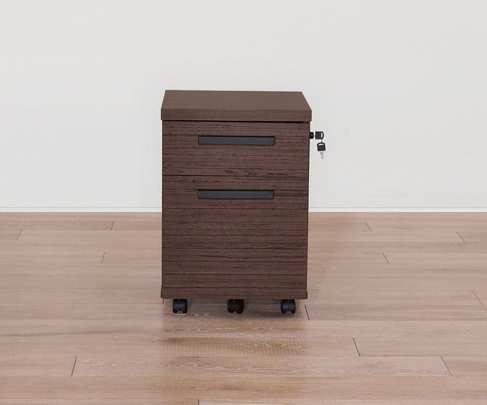 The Cary Mobile Pedestal is a brown wooden filing cabinet with a sleek walnut finish, featuring two drawers, black handles, a lock with key, and four wheels for easy mobility. This versatile office storage solution is placed on a light wood floor against a plain white wall.