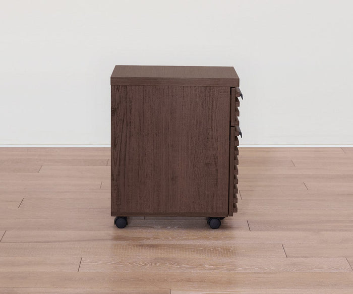 The Cary Mobile Pedestal, featuring a brown walnut finish, is positioned on a light wood floor against a plain white wall. Shown from the side with its handles and drawers out of view, this understated design offers a practical office storage solution.