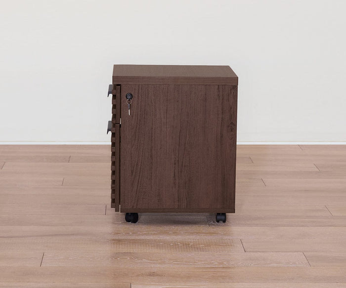 The Cary Mobile Pedestal—a wooden filing cabinet with a lock and key, finished in a rich walnut hue—features three spacious drawers and four black caster wheels. It is placed on a light-colored wooden floor against a plain white wall, making it perfect for stylish office storage.