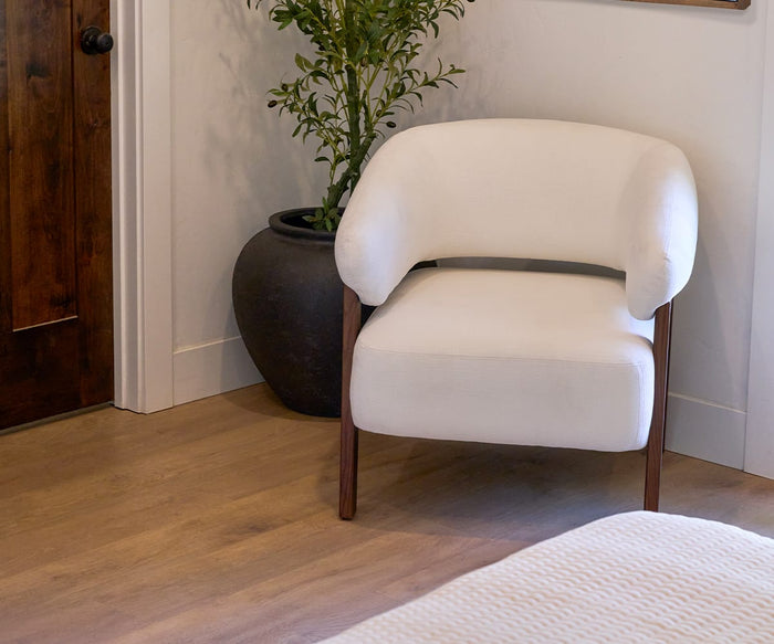 A modern white upholstered chair with wooden legs sits beside the Coleen Planter—an expansive black fiberstone piece filled with greenery—on a light wooden floor near a closed wooden door. Part of a bed with a textured white cover is visible.