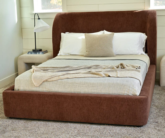 A brown upholstered bed with beige bedding and three pillows—topped with the cozy Cooper Throw, crafted from cotton yarn—stands on a carpeted floor between two small tables, one holding a lamp and books, in a softly lit bedroom.
