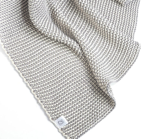 A close-up of a textured, knitted Cooper Throw in the Cooper Fog color, crafted from soft cotton yarn with a small fabric tag in one corner, displayed against a white background for a retro-cozy touch.