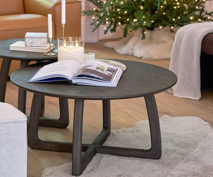 A round dark wood coffee table with an open book, candle, and matches on top sits in a retro-cozy living room adorned with a Christmas tree, flickering candles, and a beige rug. The inviting space is completed by the soft texture of the Cooper Throw crafted from cotton yarn, adding warmth and comfort to the scene.