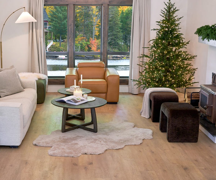 A cozy living room with a Christmas tree, string lights, a sofa draped in a Cooper Throw, an armchair, coffee table, and a large window overlooking a lake and autumn trees. A faux fur rug and candles add warmth to this retro-cozy scene.
