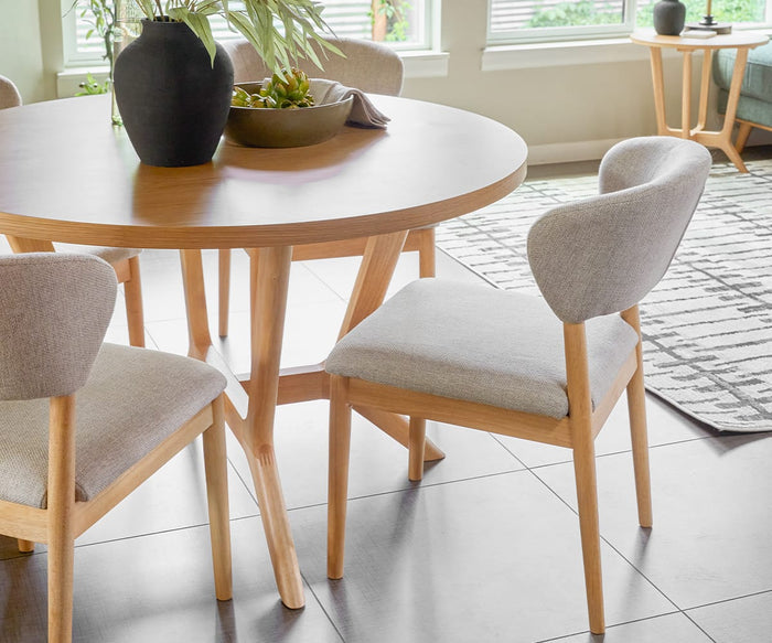 A round wooden dining table is paired with Courbe Dining Chairs, featuring Scandi-inspired light gray upholstery and crafted from sustainable rubberwood, set atop a tiled floor. The table is accented with a black vase filled with greenery and a bowl of fruit. Large windows allow natural light to fill the room.