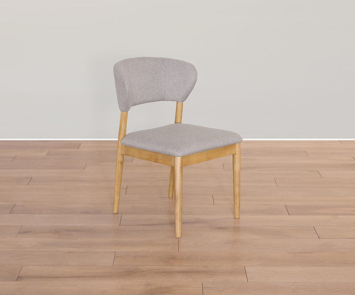 The Courbe Dining Chair, inspired by Scandinavian design and crafted from sustainable rubberwood, features an upholstered seat and backrest in light gray. It is elegantly positioned on a light brown wooden floor with a plain, light-colored wall as its backdrop.