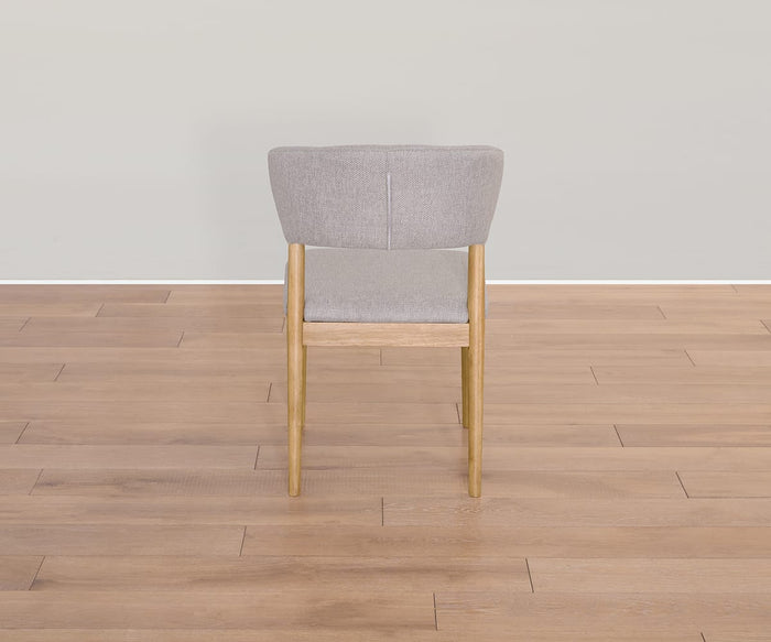 The Courbe Dining Chair, inspired by Scandinavian design and crafted from sustainable rubberwood, showcases a light wood frame paired with a gray upholstered seat and backrest. It is presented from the rear on a light brown wooden floor against a plain light-gray wall.