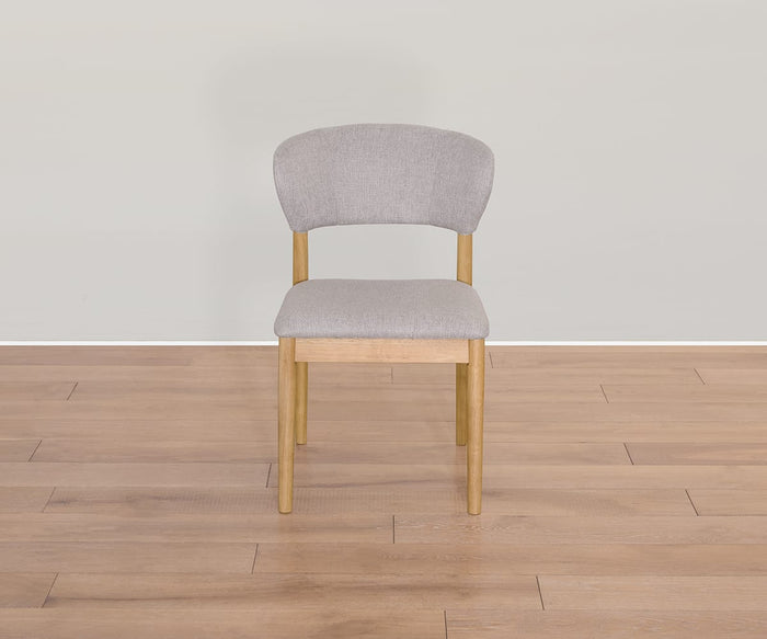 The Courbe Dining Chair, featuring Scandi-inspired wooden legs and backrest, is upholstered in light gray fabric and crafted from sustainable rubberwood for an eco-friendly touch. It sits on a light wood floor against a plain, pale wall.