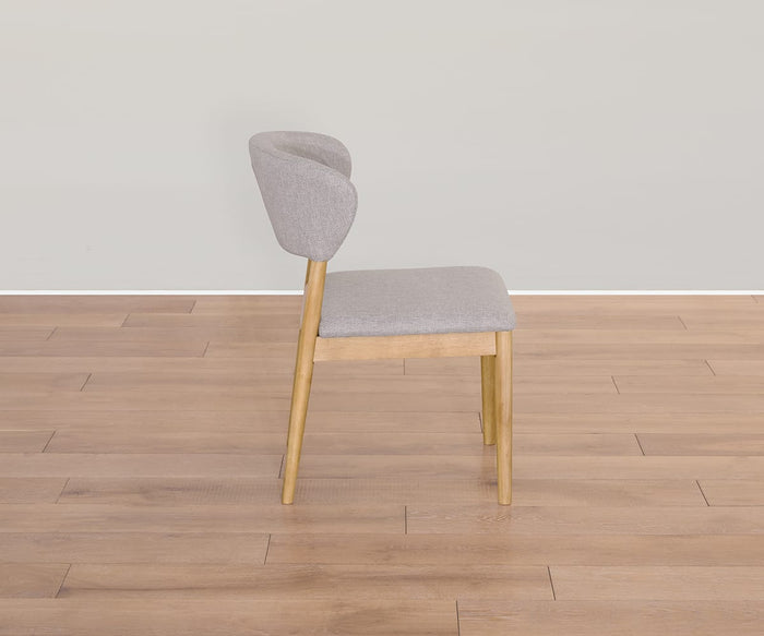 The Courbe Dining Chair, shown in a side view and set on a light brown wooden floor against a plain light gray wall, features Scandi-inspired design with clean lines. Crafted from sustainable rubberwood and upholstered in light gray fabric, this modern chair highlights natural materials.
