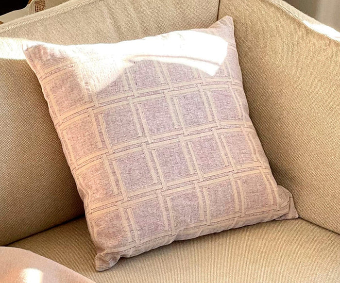 A light-colored Fair Trade Dahlia Cushion Cover featuring a geometric square pattern rests on a beige fabric sofa, its organic cotton softly illuminated as sunlight casts a gentle shadow across the pillow.