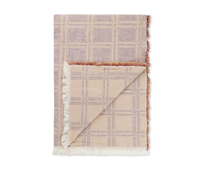 The Dahlia Throw, shown folded with a corner flipped to reveal both sides of its light plum grid design, is crafted from organic cotton and finished with fringed edges. Fair Trade and featuring a textured weave, this throw adds depth and softness to any space.