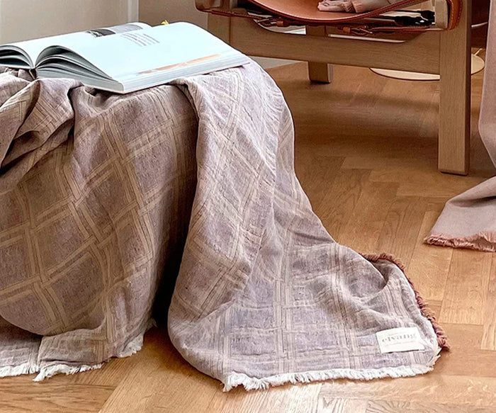 A Dahlia Throw with a textured weave drapes over the edge of a wooden table, partially covering an open book. In the background, a wooden chair with a folded Dahlia Throw made from organic cotton is set on a light wood floor.