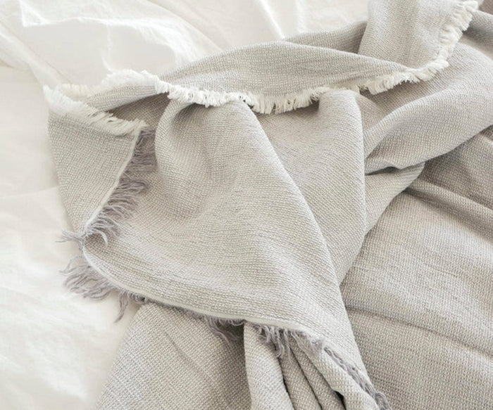 The Daisy Bedcover, featuring a light gray hue, textured organic cotton fabric, and fringed edges, is draped across a white bed to create a cozy and inviting appearance.