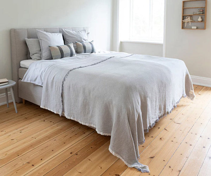 A neatly made bed featuring the Daisy Bedcover in light gray, adorned with multiple pillows, sits in a bright room with wooden floors, a small stool, and shelves on the wall near a window.