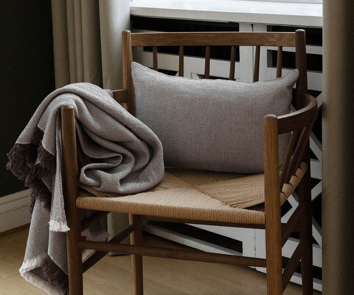A wooden chair with a woven seat holds a gray rectangular Daisy Cushion Cover and a gray fringed blanket draped over one arm, positioned next to a window with white curtains.