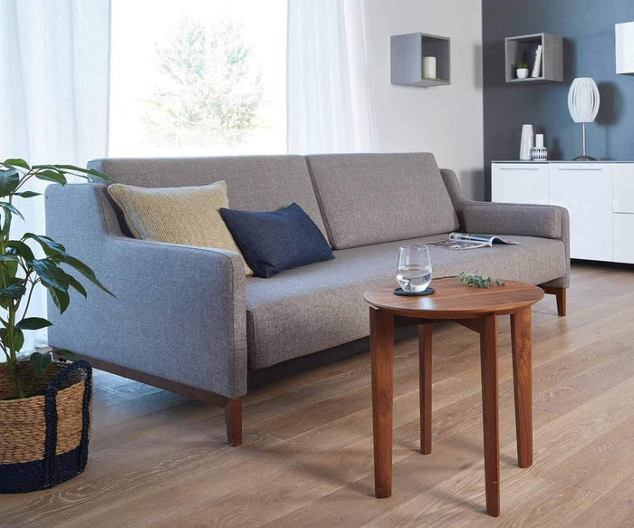 A modern living room features a gray sofa with two pillows, a round wooden side table holding water and a plant, a potted plant, and minimalist white shelves and cabinets against a gray accent wall with a subtle faux-cement finish. Danne Wall Cubes offer eco-friendly storage while adding visual interest to the clean design.
