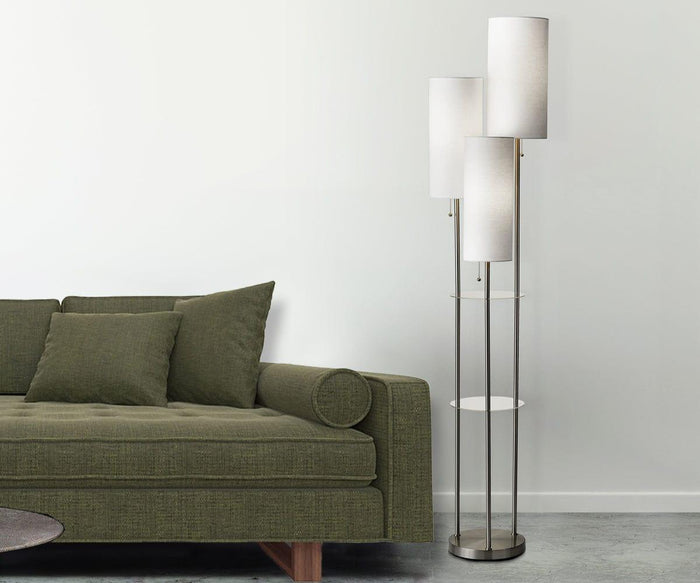 A modern olive-green sofa with cushions sits beside the Daphne Shelf Floor Lamp, which boasts a tall, three-shade silhouette, metallic base, and a small glass shelf, all set against a white wall and gray floor.