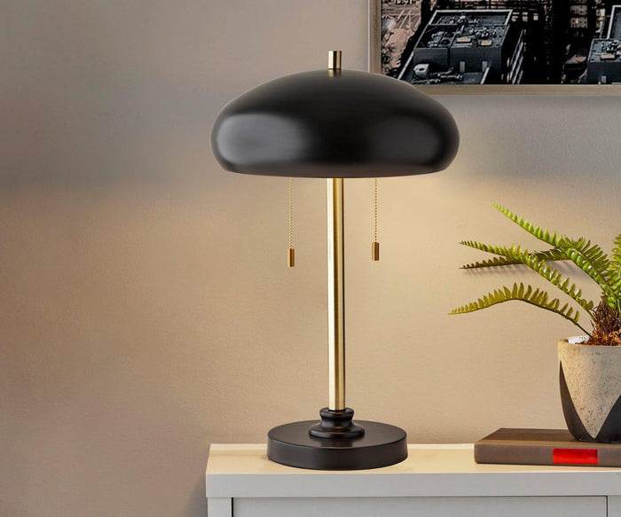 The Darius Table Lamp, featuring a black and gold finish and a dome-shaped metal shade, sits on a white table beside a potted fern and a closed red book, softly illuminating the beige wall and part of a framed city photo above.