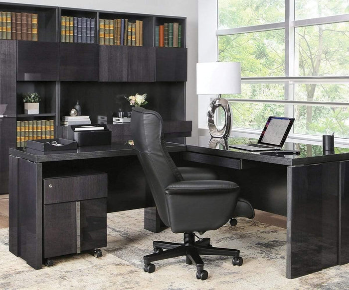 Modern office with a black L-shaped desk, black leather chair, the Darlene Table Lamp, laptop, bookshelves with books, and large windows showing greenery outside. Light rug covers the floor.