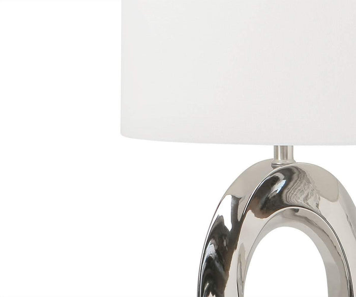 The Darlene Table Lamp showcases a sleek, silver oval base with a modern sculptural design, complemented by a simple white lampshade and displayed on a crisp white background.
