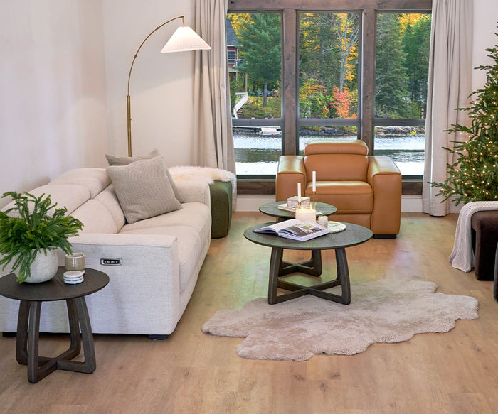 Modern living room with a beige sofa, tan armchair, round coffee tables, a potted plant, and a lit Christmas tree. The Delaney Arc Lamp serves as a statement piece beside the seating area. Large windows show a lake and trees with autumn colors outside.