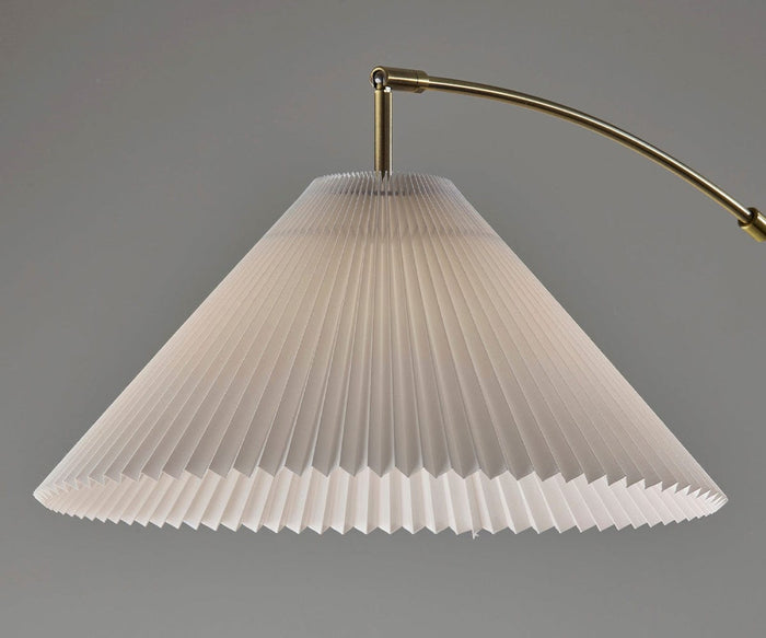 A close-up of the Delaney Arc Lamp showcases its pleated, cone-shaped white lampshade paired with an antique brass arm, set against a plain gray background—an understated statement piece that exudes timeless elegance.