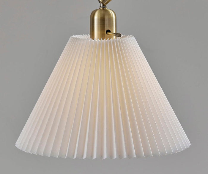 The Delaney Table Lamp, featuring a brass base and a pleated white lampshade, is gently illuminated against a plain grey background, evoking an air of elegance.