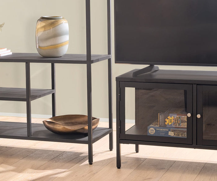 A black metal TV stand holds a TV and board game, next to the modern Heroy 48″ Bookcase displaying a decorative vase and wooden bowl, all set on a light wooden floor.
