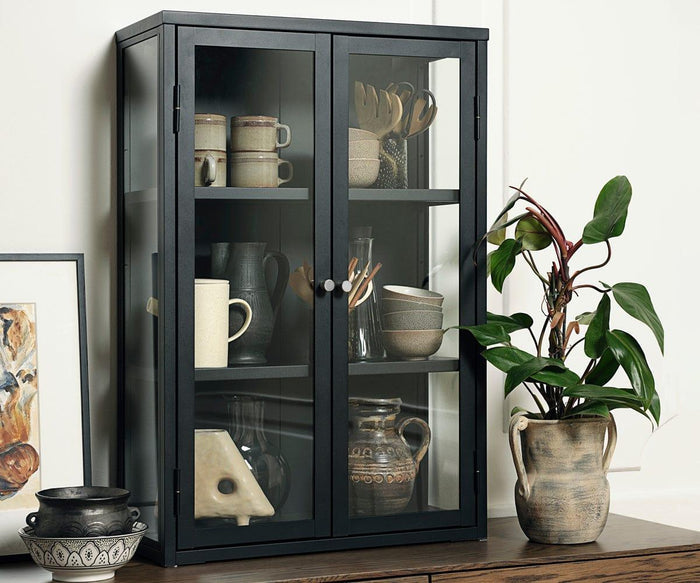 The Dion Tall Wall Cabinet, featuring a sleek black glass front, stores ceramic dishes and cups while adding contemporary style to the room. Beside it, a potted plant in a rustic vase, a framed picture, and pottery are arranged on a white surface against a light-colored wall.
