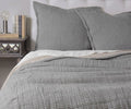 A neatly made bed with light gray textured bedding and pillows, highlighted by a Dorian Sham in Dorian Fog that features Kantha-style stitching and is crafted from 100% cotton, sits next to a nightstand with books, a lamp, and a mirrored drawer. The wall and headboard are light-colored.