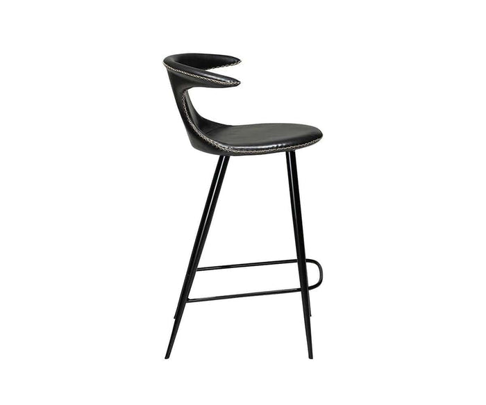 The Drake Counter Stool features a black, modern silhouette with a curved backrest, slim metal legs, and an integrated footrest. Its seat and back are upholstered in chic faux leather with decorative edge stitching, showcasing modern Danish design influences. The stool is presented against a white background.