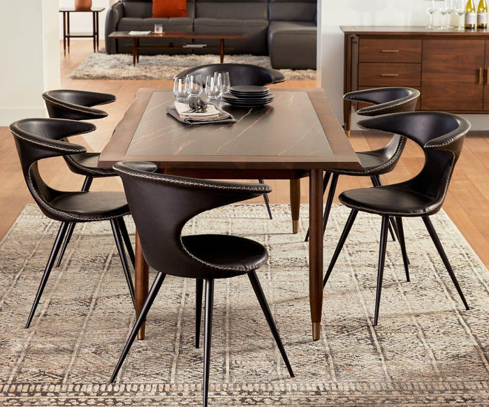 A modern dining room with a wooden table surrounded by six Drake Dining Chairs—black faux leather seats with metal legs. Plates, glasses, and utensils are set on the table, which rests on a patterned rug, while a sideboard and sofa appear in the background.