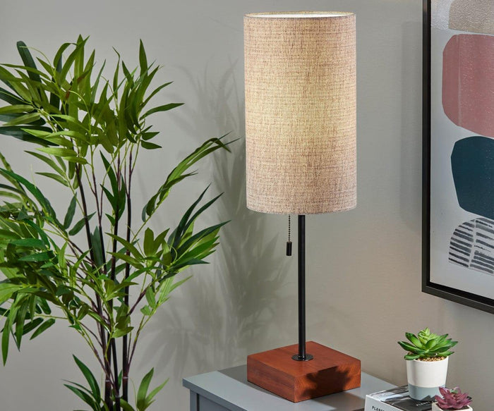 The Dusk Table Lamp, featuring a tall cylindrical design with a beige shade and walnut base, sits on a gray table next to a green plant. A partial view of abstract art and a small succulent are also visible, enhancing the mid-century decor vibe.