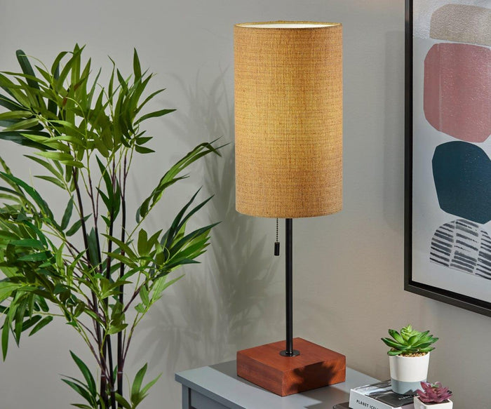 The Dusk Table Lamp, featuring a walnut base and a beige cylindrical shade, sits on a gray surface beside a green potted plant, framed abstract artwork, and small succulents, bringing mid-century decor charm to the setting.
