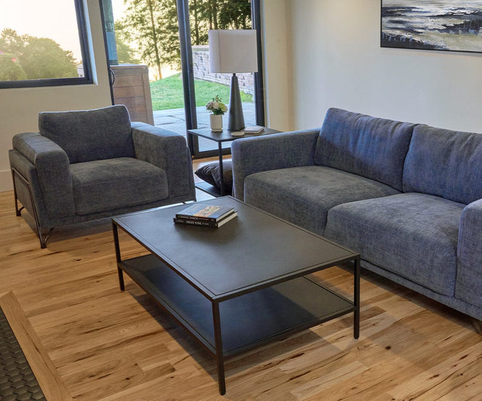 Modern living room with a gray sofa, matching armchair, black cement coffee table topped with books, side table displaying a plant, wood flooring, large windows, and a painting on the wall. The Dyre Table Lamp adds a weather-resistant accent to the space, seamlessly blending style and practicality.