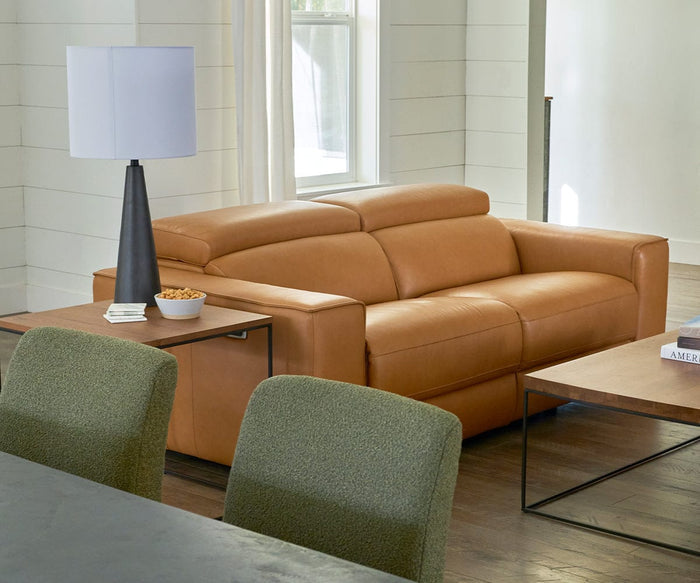 A light brown leather sofa sits in a living room with a side table holding the Dyre Table Lamp, snacks, and a remote. Green chairs and a weather-resistant wooden coffee table are also visible as sunlight streams through the window.