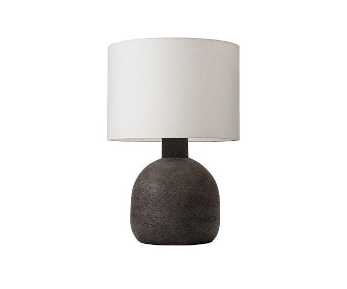 The Earthenware Jug Lamp features a rounded dusty black terracotta base reminiscent of traditional earthenware, paired with a large white cylindrical lampshade, set against a plain white background.
