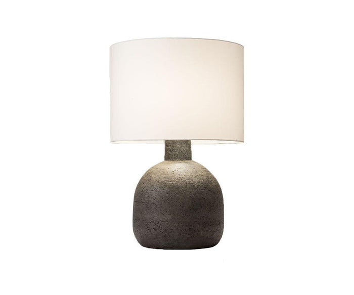 The Earthenware Jug Lamp is a modern table lamp featuring a round, textured gray concrete base inspired by an earthenware jug and topped with a large, white cylindrical lampshade, shown against a plain white background.