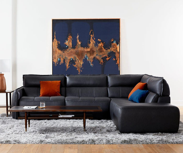 A modern black sectional sofa with orange and blue pillows sits on a gray rug. A wooden coffee table with books and a glass is in front, complemented by the Earthy Luster Lamp with its matte-finished base that adds warmth to the scene. An abstract artwork with blue and copper tones hangs on the white wall behind.