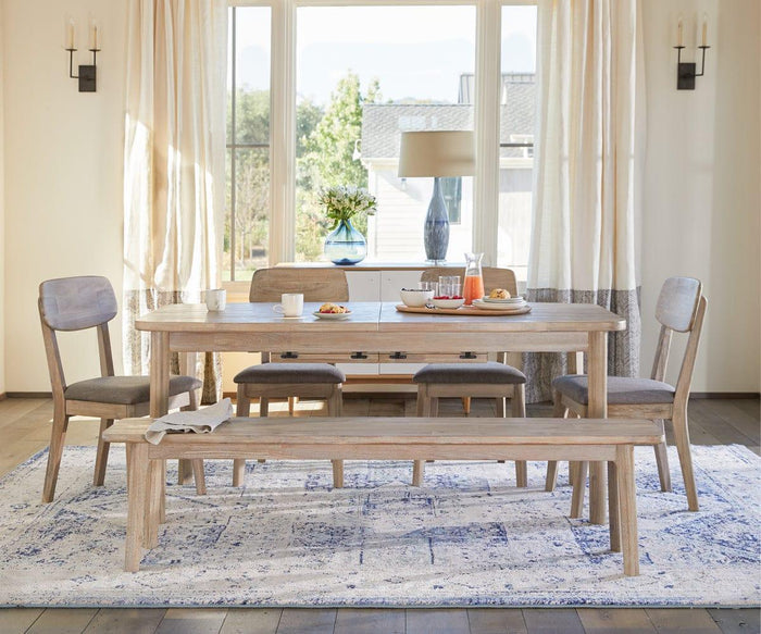 The Eckler Bench, crafted from solid acacia wood, pairs with a wooden dining table and four chairs atop a blue and white rug. Breakfast items and cups rest on the tabletop, while sunlight pours in through large curtained windows.