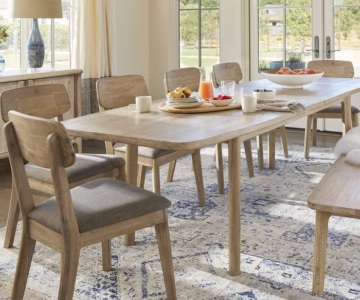 A wooden dining table with six Eckler Dining Chairs—each featuring a comfortable upholstered seat—is set for breakfast with plates, cups, orange juice, pastries, and fruit on a blue-patterned rug in a bright room with large windows.