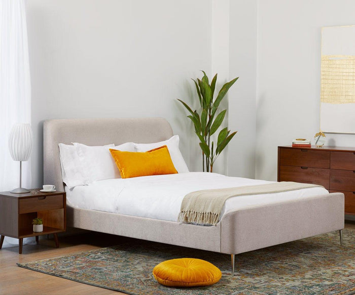 A modern bedroom features a beige upholstered bed with crisp white bedding and an orange accent pillow. On the rug sits a round orange cushion, while a plant, wooden nightstand, dresser, and abstract wall art provide a stylish backdrop. The nightstand is enhanced by the Egg Table Lamp, showcasing its white pleated shade and satin steel base for a sleek contemporary touch.