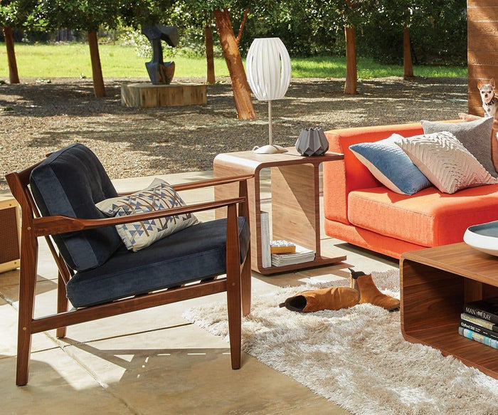 Mid-century modern patio featuring a navy chair, orange couch, side table, rug, and pillows. The area is illuminated by the Egg Table Lamp with its satin steel base and white pleated shade. Sunlight filters into the outdoor setting adorned with trees and greenery in the background.