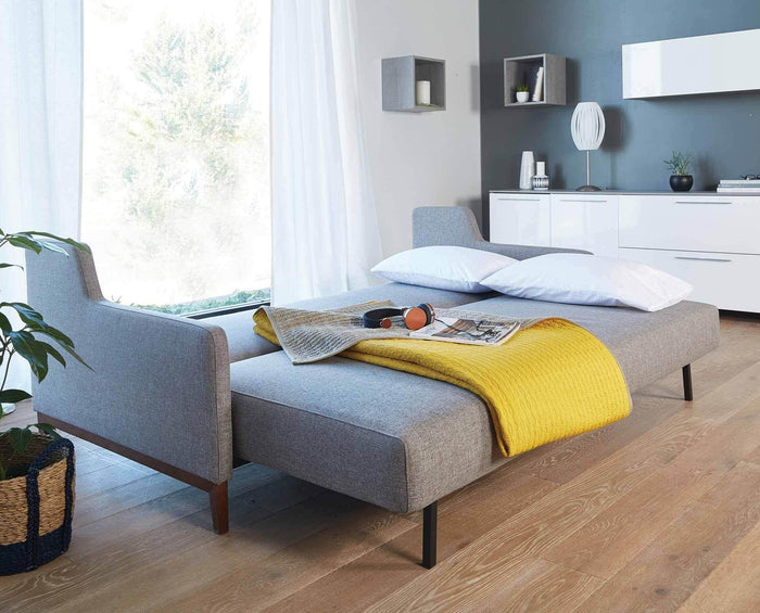 A sleek gray sofa bed adorned with white pillows and a yellow blanket is arranged in a sunlit room with wood flooring, a large window, potted plant, and simple wall décor. In the background, a dresser is highlighted by the Egg Table Lamp, which features a satin steel base and a white pleated shade.
